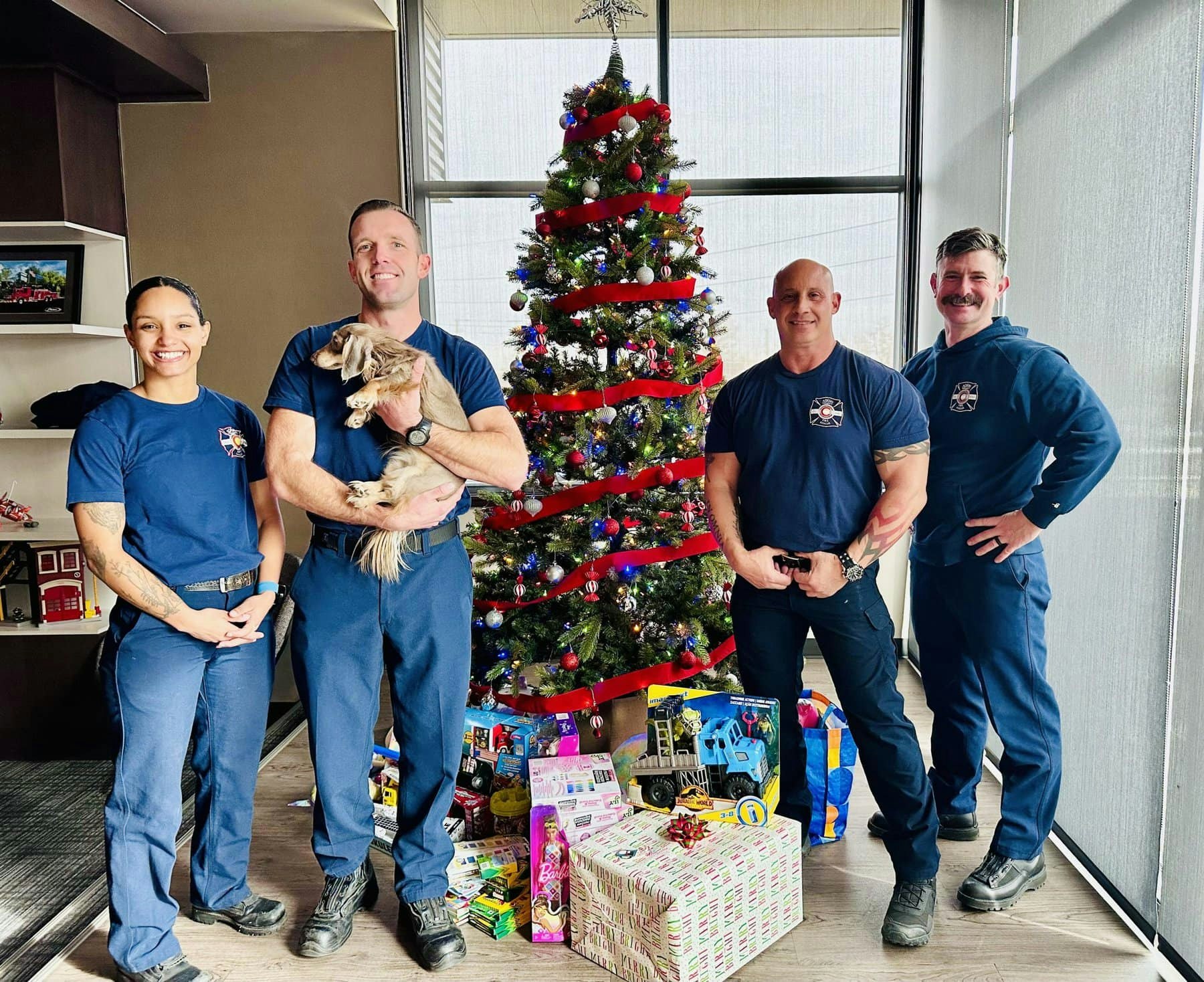 Four people in uniform with a dog, standing by a decorated Christmas tree and gifts.