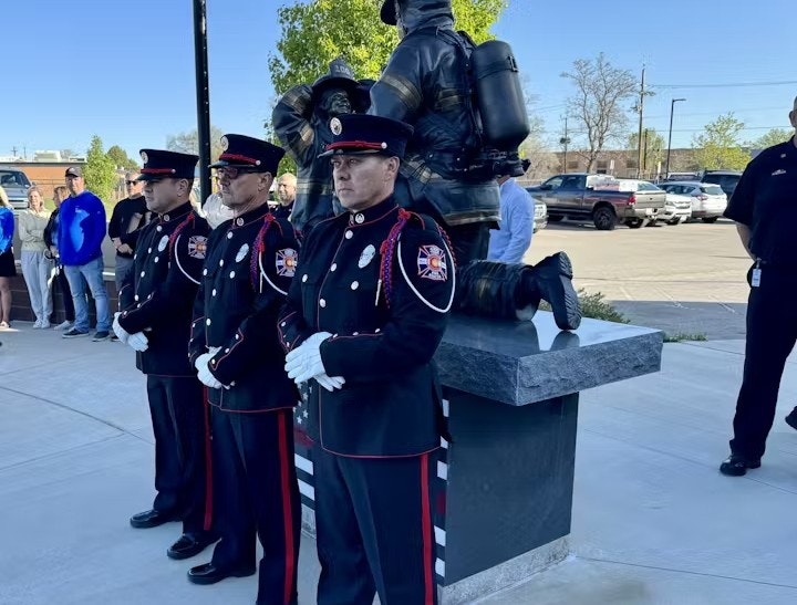 Three firefighters in formal uniforms stand beside a firefighter statue, with onlookers nearby.