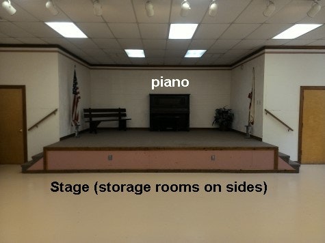 inside updated community building showing new floor to original stage with piano in background.  Storage rooms on each side.