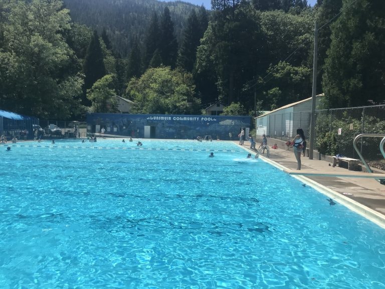 Looking down the length of the swimming pool bath house in far end with trees and mountain beyond that. A few in pool and on deck.