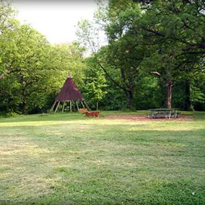 A grassy area surrounded by trees, featuring a teepee structure and a fire pit with benches nearby.
