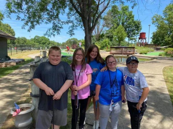 A group of five children poses for a photo in a park on a sunny day, smiling and enjoying their time together.