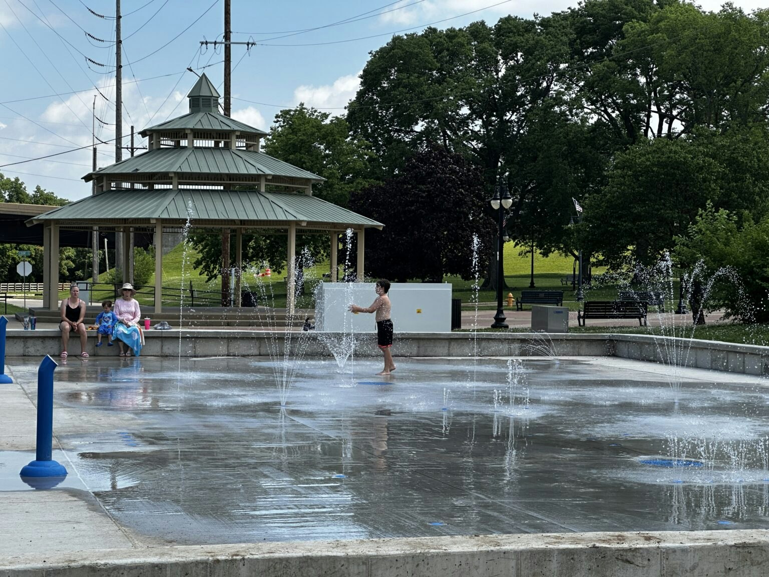 A child plays in a splash pad while others relax nearby under a gazebo in a park on a sunny day.