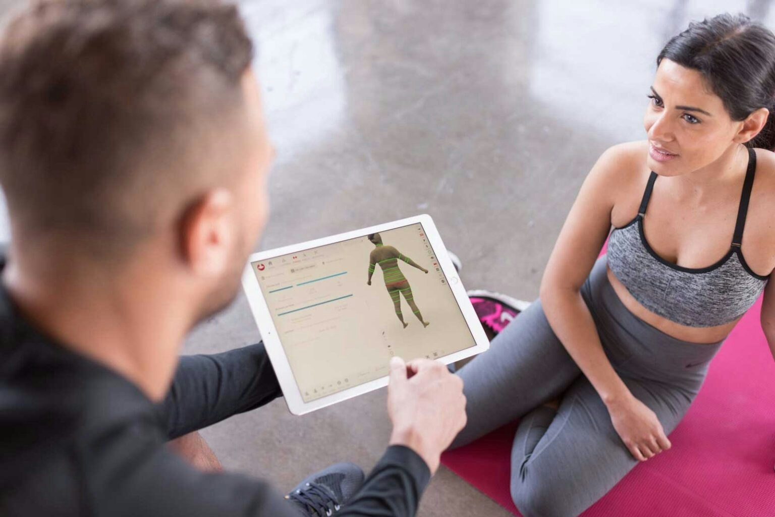 A trainer shows a client an interactive display on a tablet, likely related to fitness or body analysis, during a workout session.