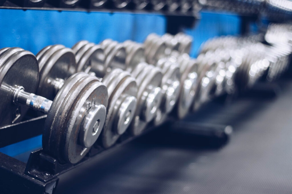 The image features a row of metal dumbbells on a rack in a gym, with a blurred blue background.
