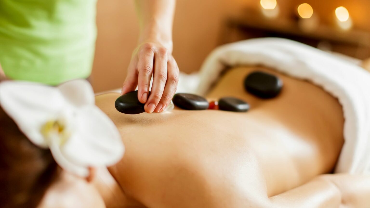 A person is receiving a hot stone massage, with smooth stones placed on their back and a flower nearby for relaxation.