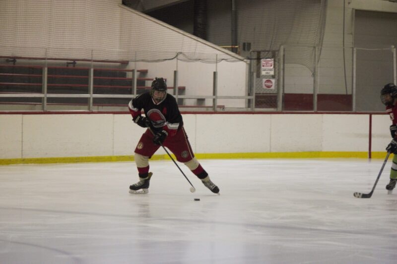 A hockey player in black and red uniform skates on ice, controlling a puck while another player is nearby.
