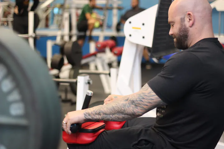A muscular man with tattoos is using gym equipment in a fitness center, focused on his workout.