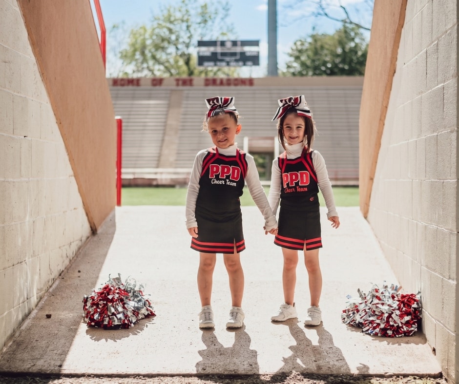 Two young cheerleaders holding hands, wearing matching outfits and bows, with pom-poms nearby in a stadium setting.