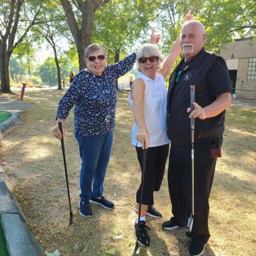 Three people are posing outdoors, smiling and holding golf clubs, enjoying a mini-golf or recreational activity.