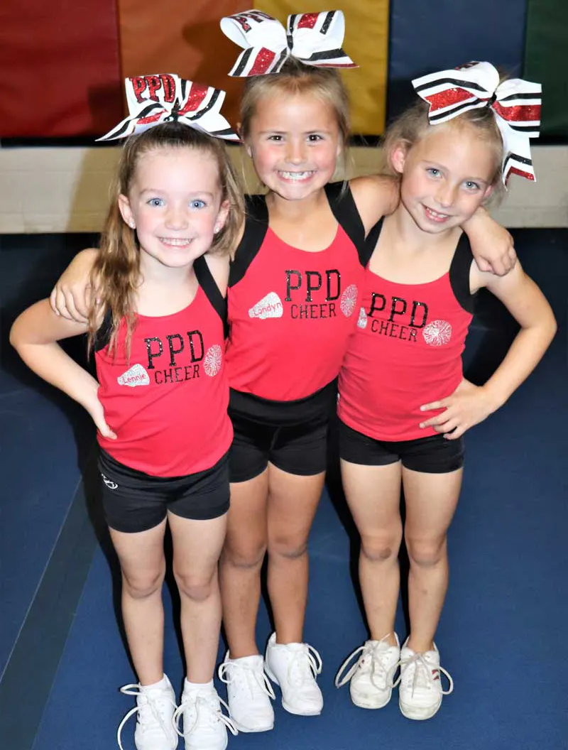 Three young cheerleaders pose together, smiling, wearing matching outfits with bows, in a gym setting.