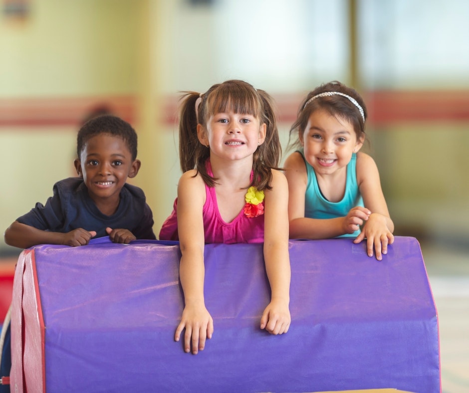 Three children, two girls and a boy, are smiling and having fun while playing on a purple gymnastics mat in a bright setting.