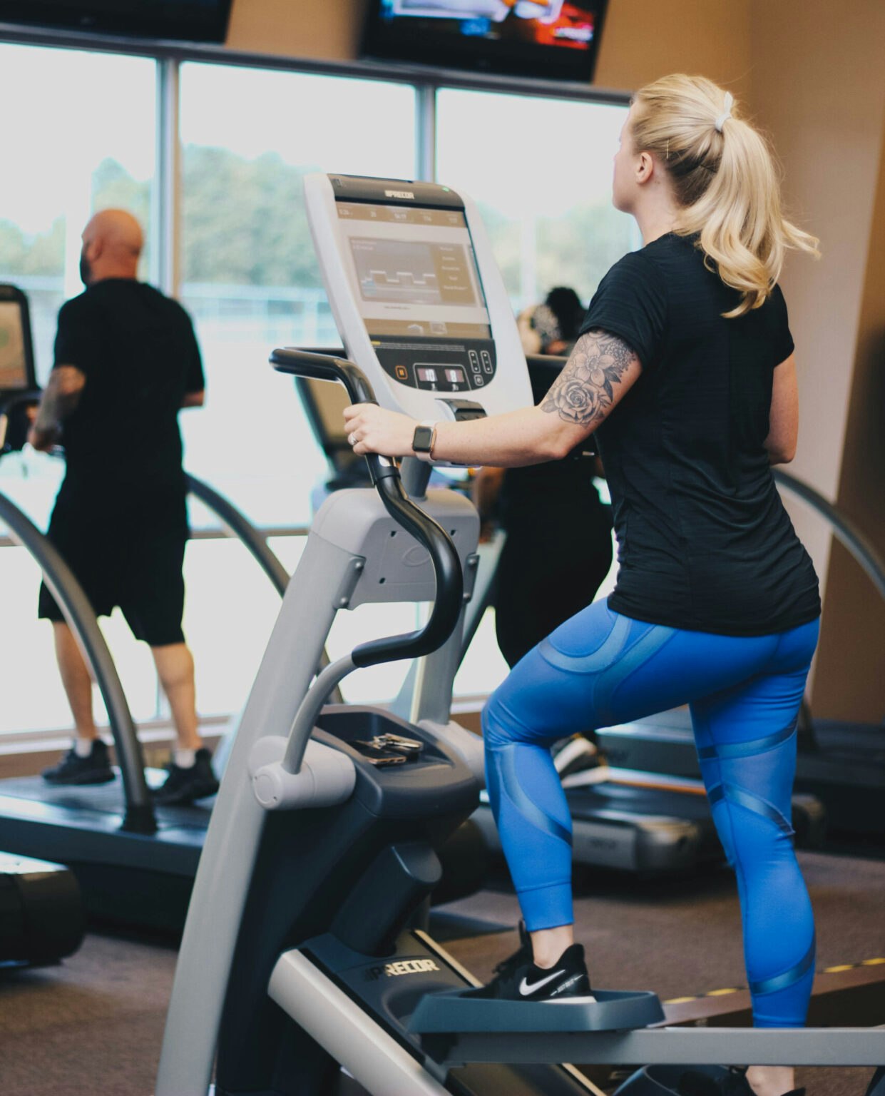 A woman exercises on an elliptical machine at a gym, with others in the background and TVs showing screens.