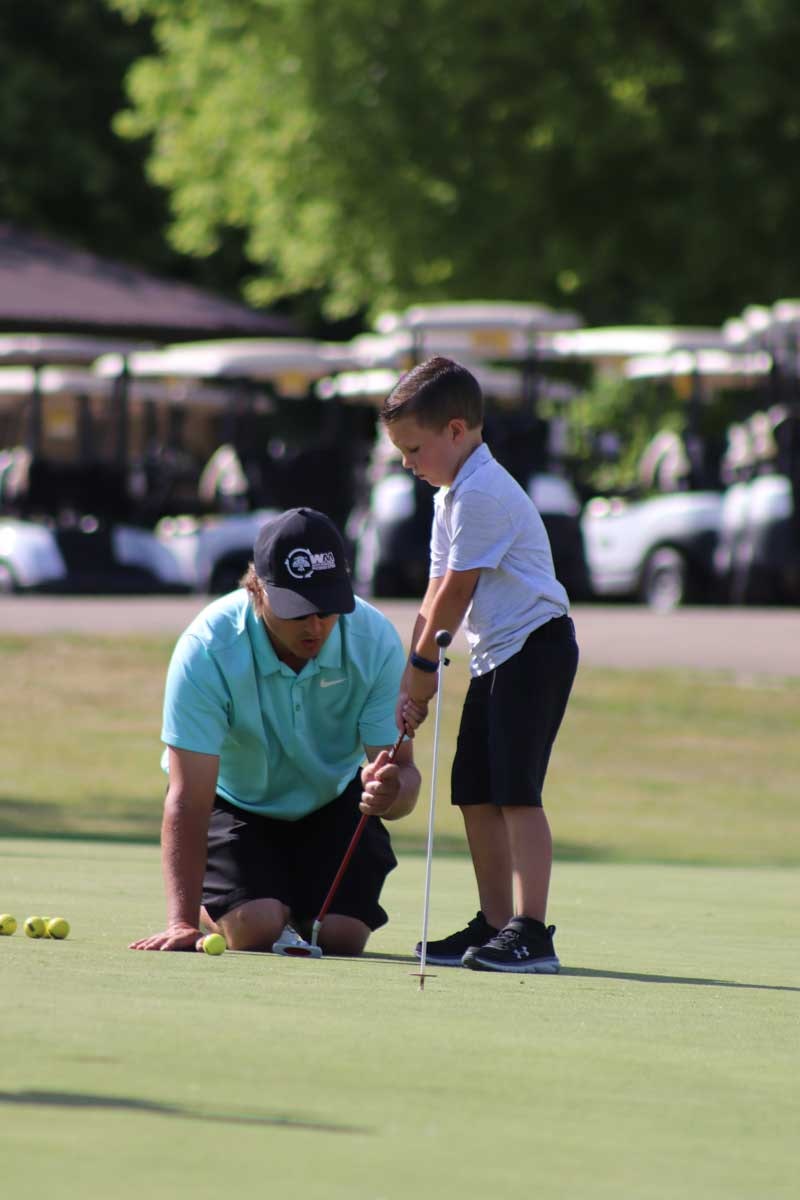 A man teaches a young boy how to putt at a golf course, surrounded by golf balls and carts in the background.