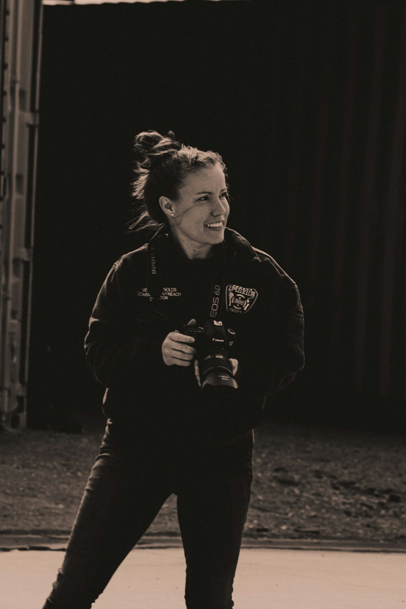 A person smiling while holding a Canon camera, wearing a dark jacket, standing outdoors.