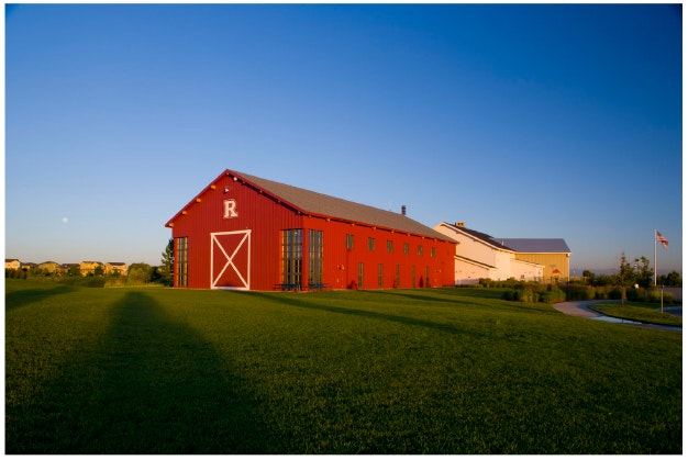 A large red barn with white trim on a green lawn under a clear sky, casting a long shadow.