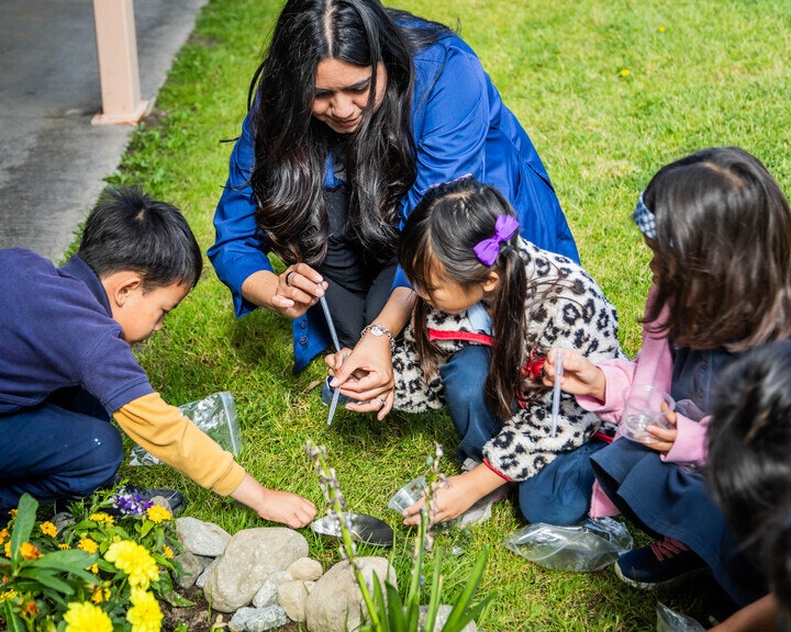 A woman and several children are engaged in a hands-on gardening or science activity outdoors, exploring nature together.