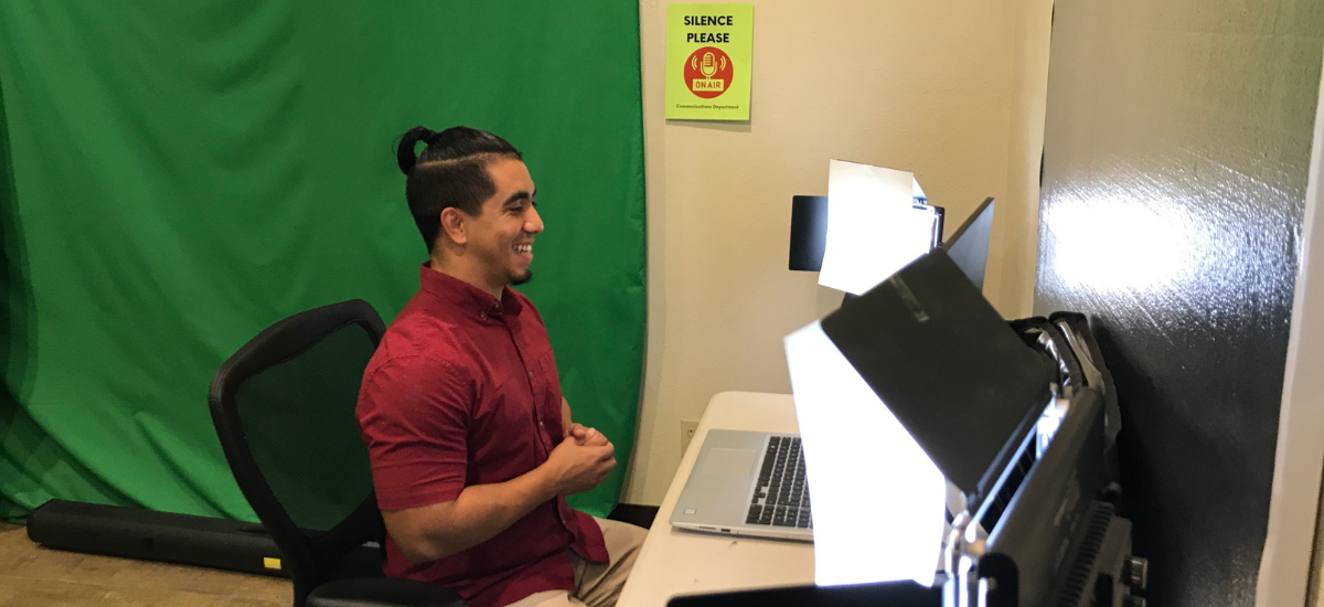 A person sits in front of a laptop with studio lights, smiling, against a green screen backdrop and a "Silence Please" sign.