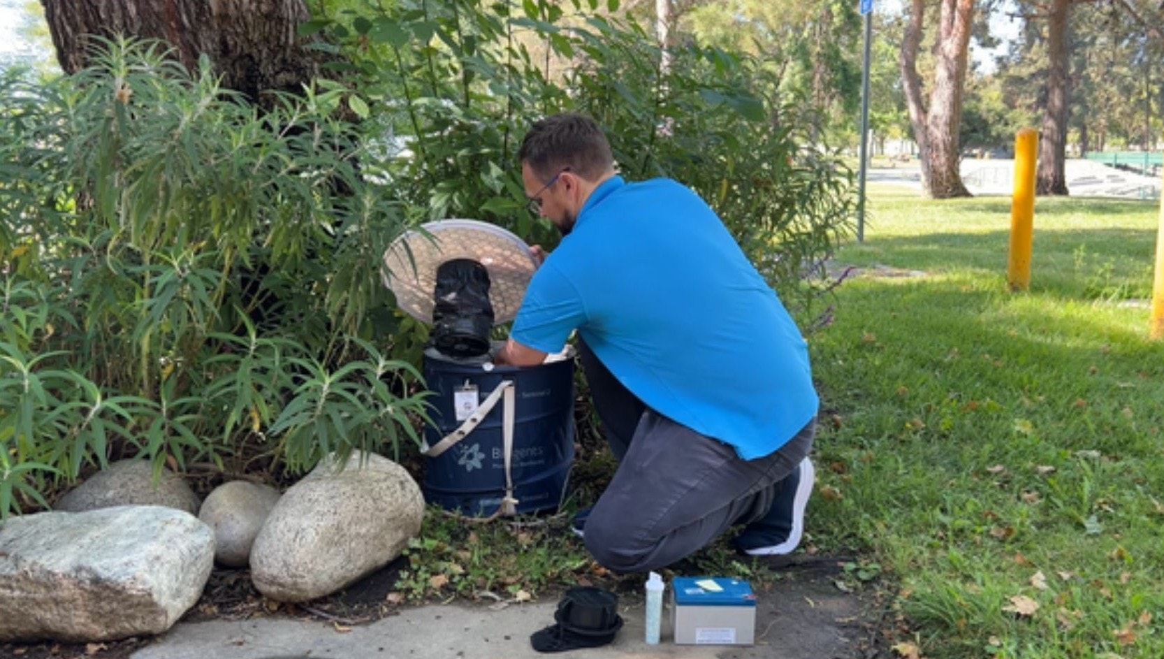 A man in a blue shirt works with a device outdoors near plants and rocks.