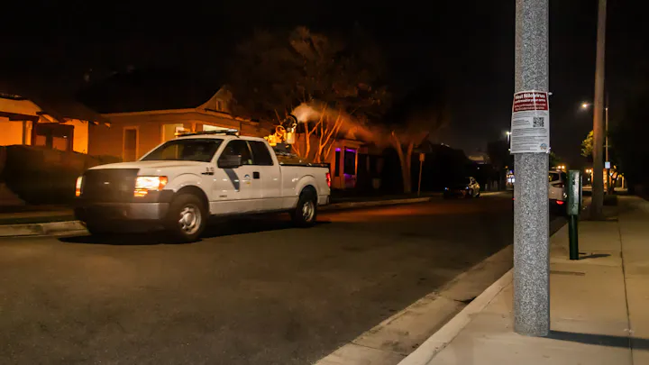 A white truck sprays pesticide on a street at night, with homes illuminated and a public alert poster about West Nile virus nearby.