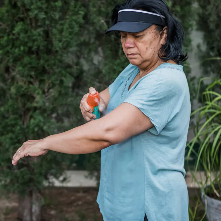 A person applying mosquito repellent to their arm while outdoors, wearing a light blue shirt and a black visor.