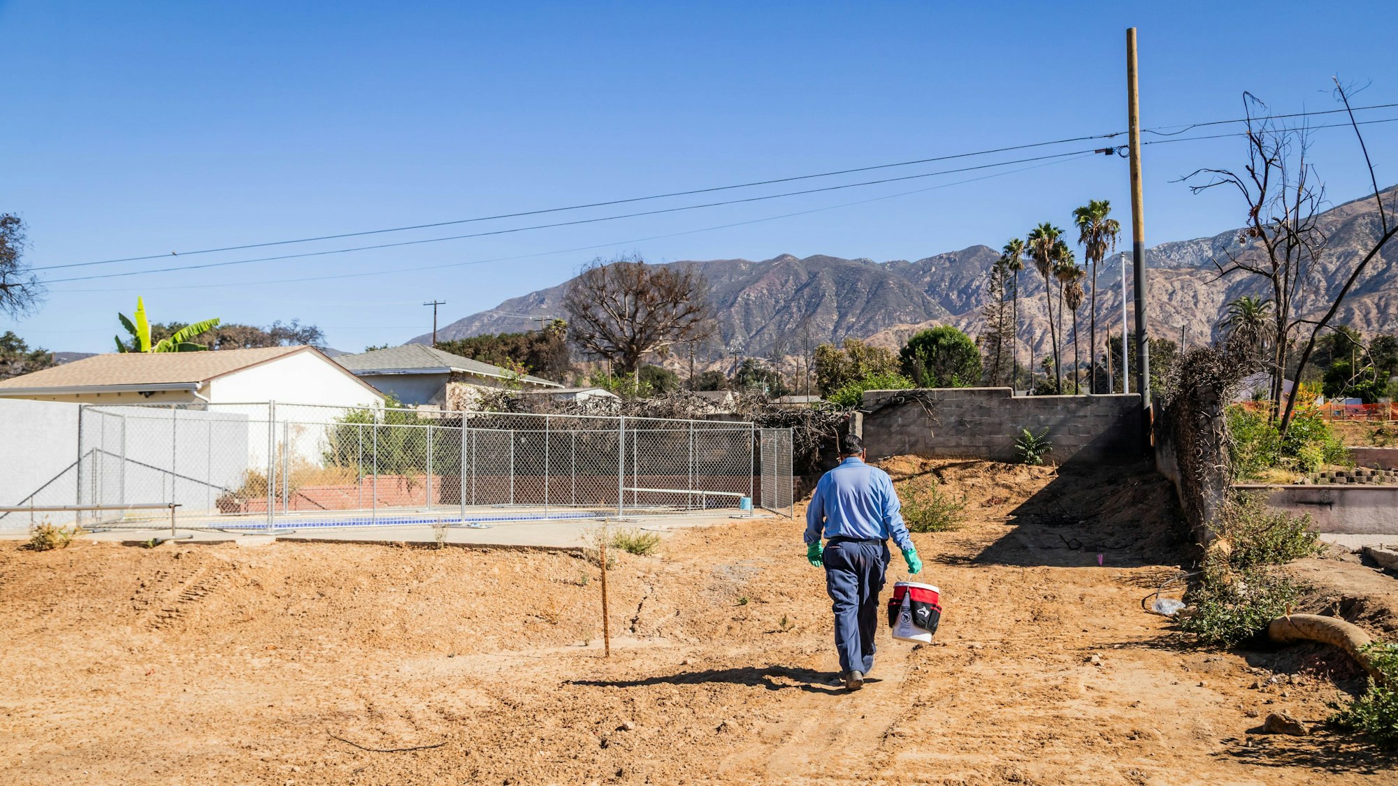 Man walking on dirt path with a bucket, wearing gloves, with mountains and trees in the background.