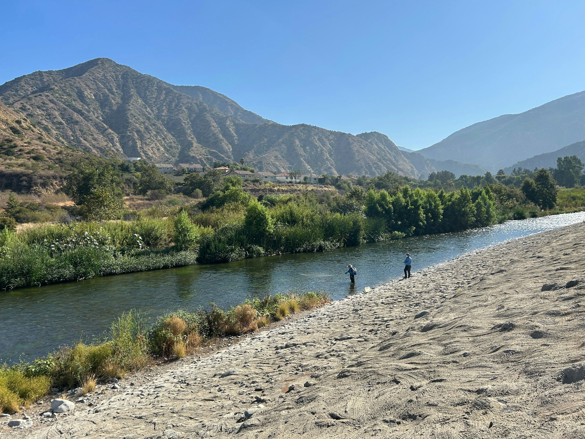 Two people wading in a river with a mountainous backdrop and lush greenery.