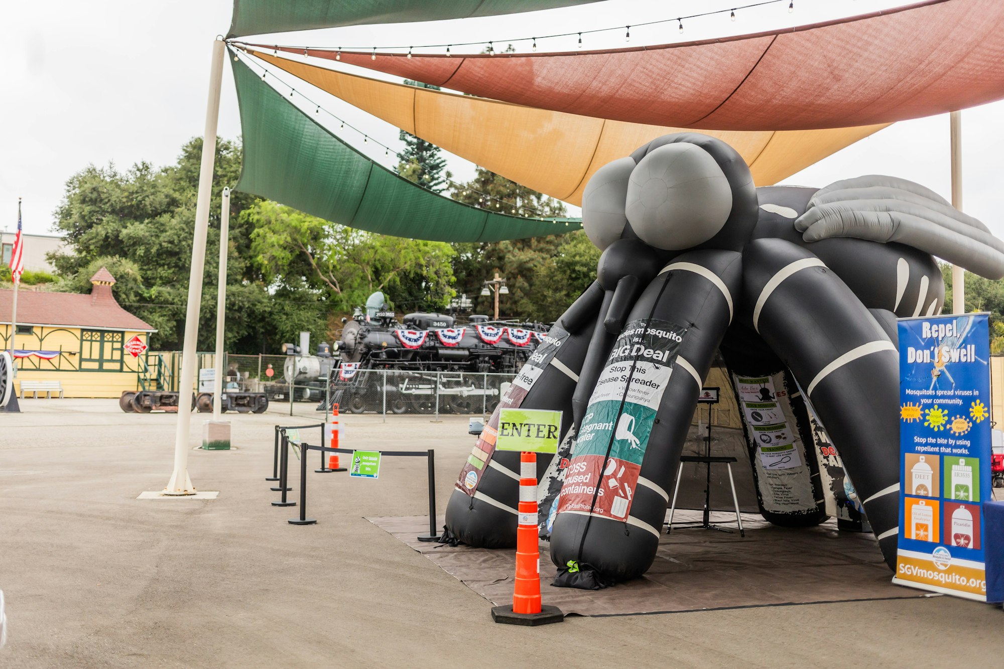 An inflatable mosquito exhibit with educational banners, a train in the background, and shaded outdoor setting.
