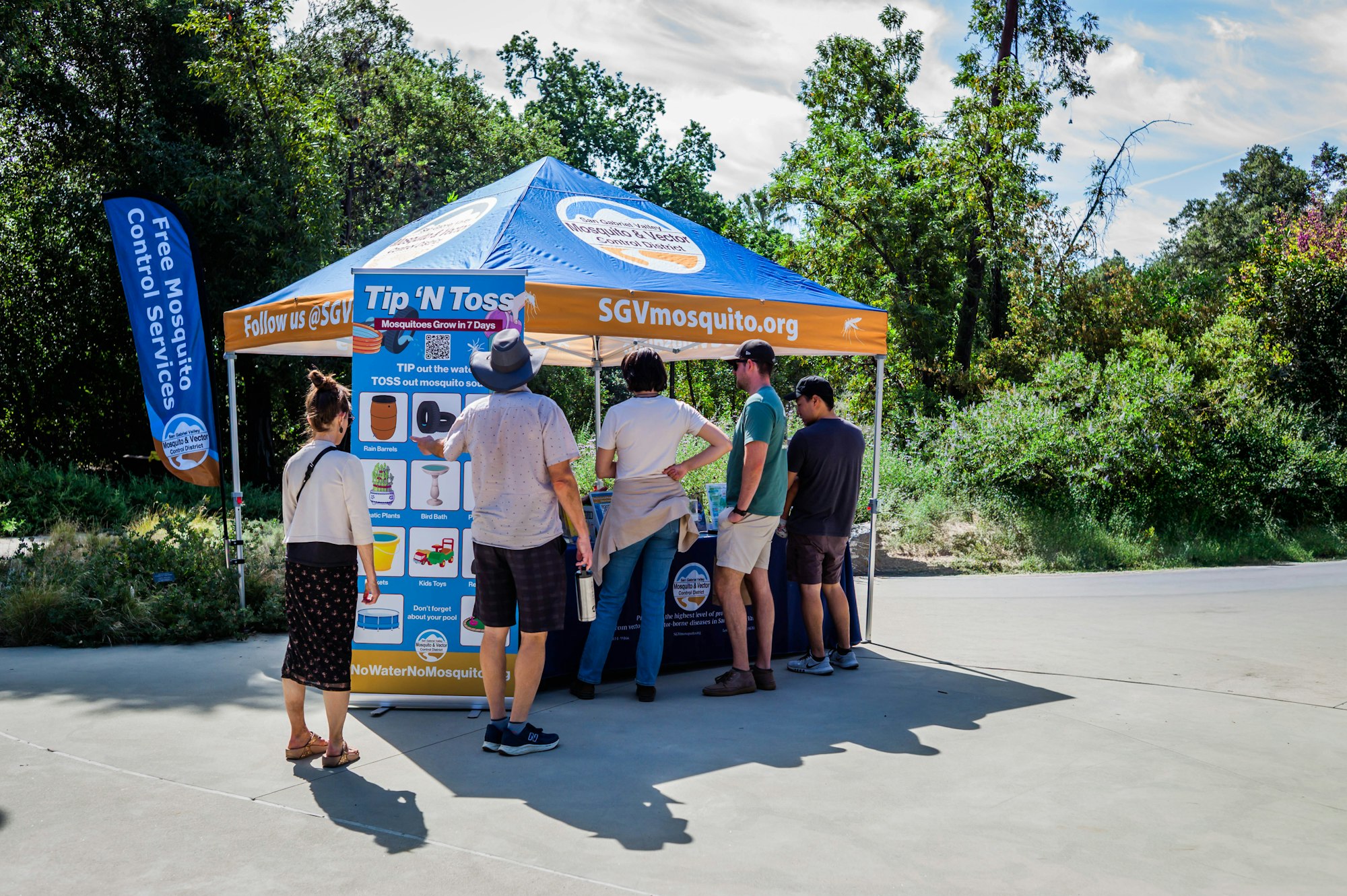 People are gathered at a tent promoting mosquito control services, featuring the "Tip 'N Toss" campaign to eliminate standing water.