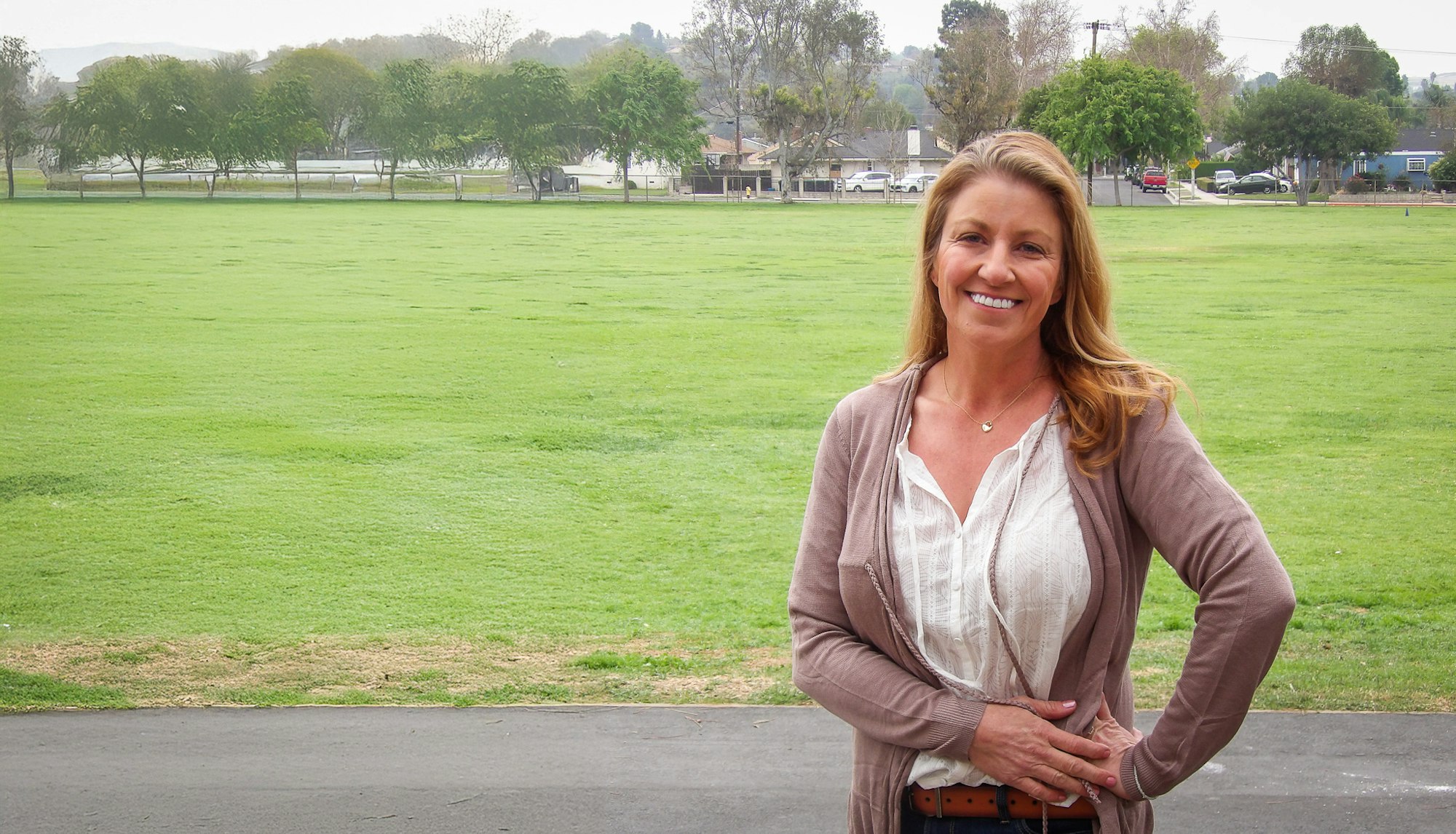 A woman smiling in front of a large grassy field with trees and a few buildings in the background.