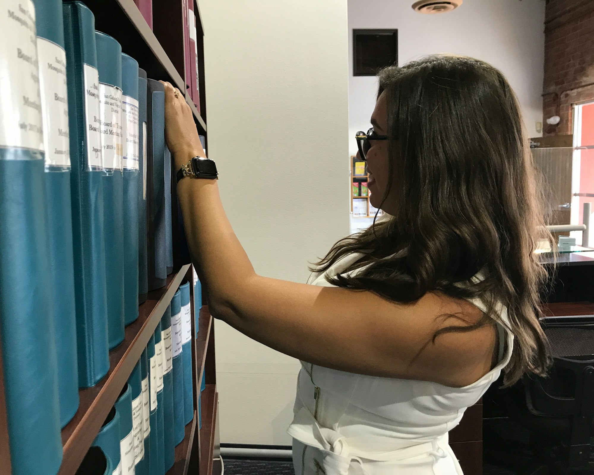 A woman in a white outfit is reaching for a blue binder on a shelf in a library or office setting.