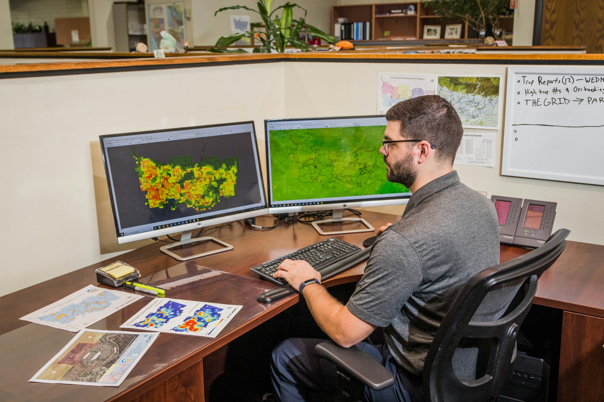 A man works at a desk with two monitors displaying data maps, with phones, papers, and a whiteboard visible.