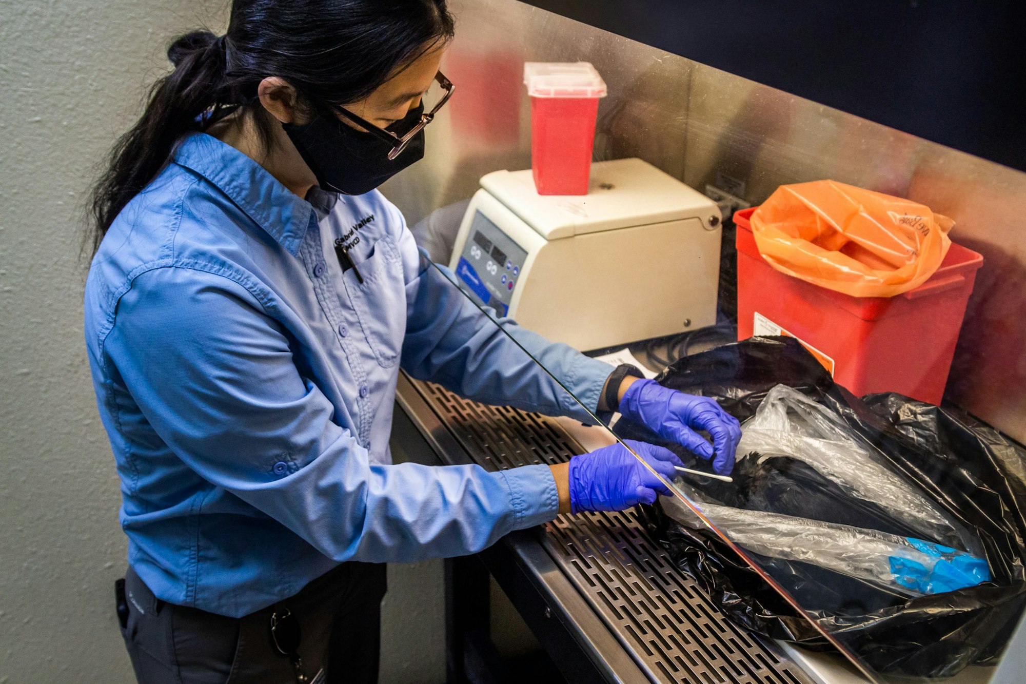 A person wearing a lab coat, gloves, and mask working with lab equipment, potentially handling samples for testing.