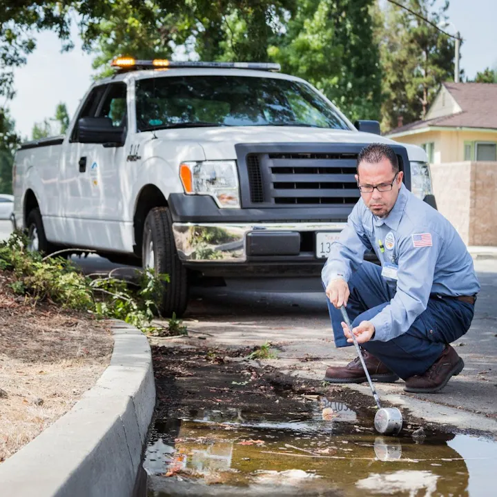 A worker in uniform inspects a puddle near a white truck, likely assessing water quality or conducting maintenance.