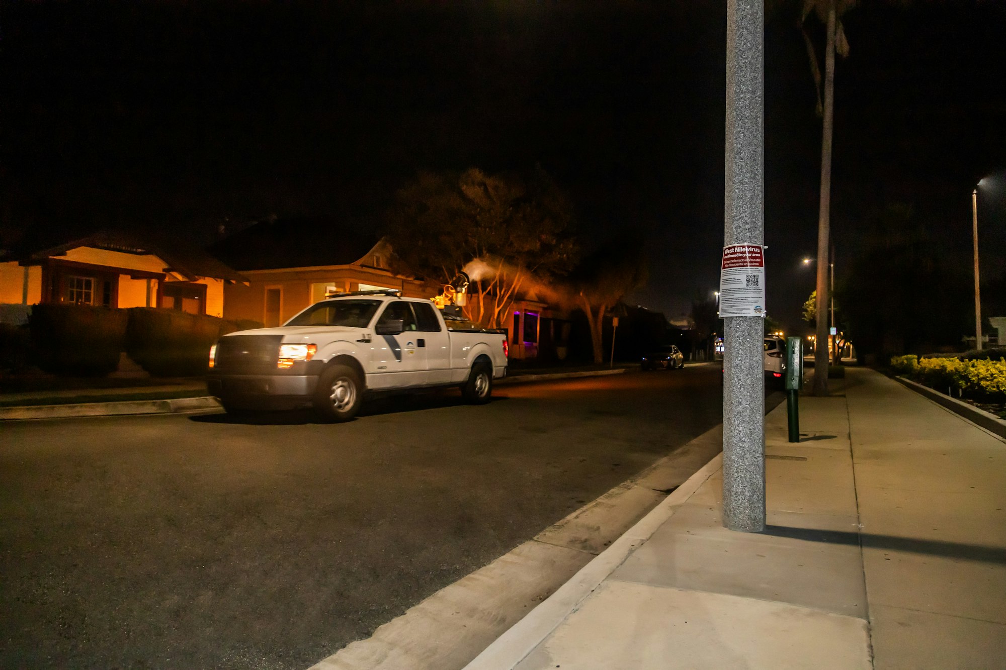 A nighttime street scene with a pickup truck, houses, streetlights, and a sign on a pole.