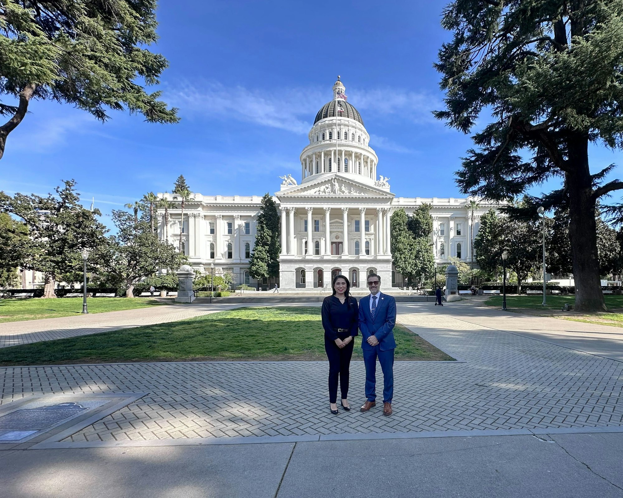 A couple stands in front of a grand government building on a sunny day, surrounded by greenery and blue skies.