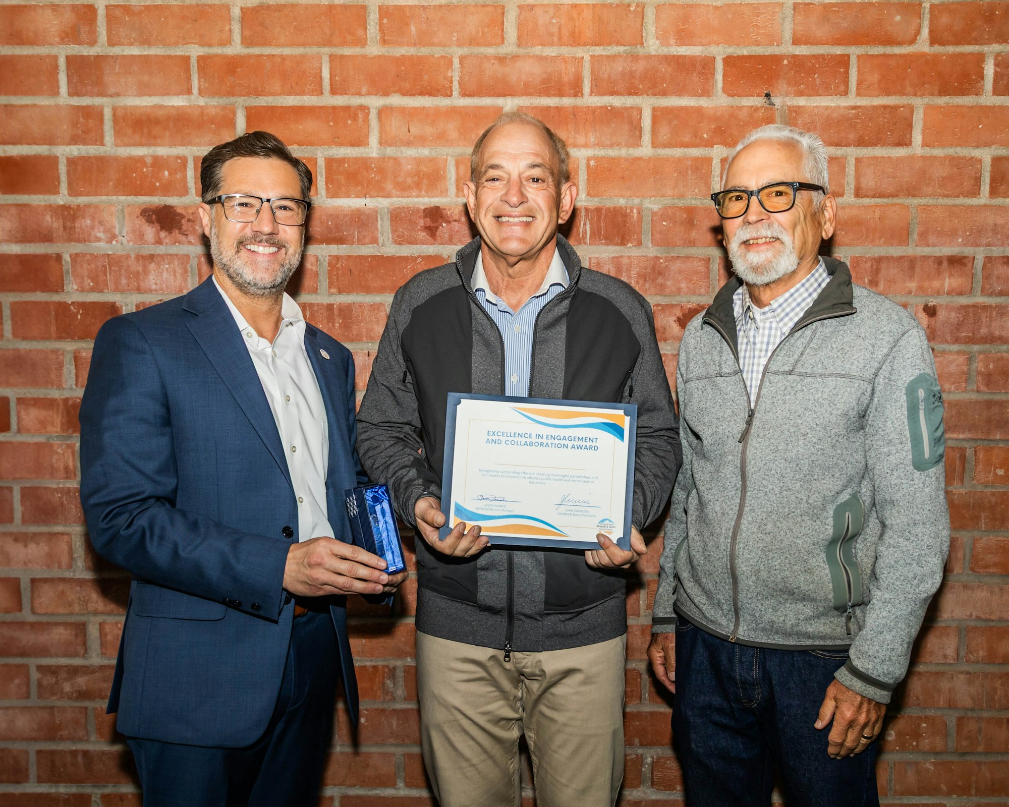 Three men standing in front of a brick wall; one holding an award certificate and another holding a blue trophy.