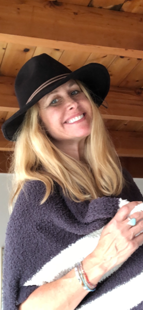 A smiling woman wearing a black hat and a cozy wrap, posed in a wooden setting.