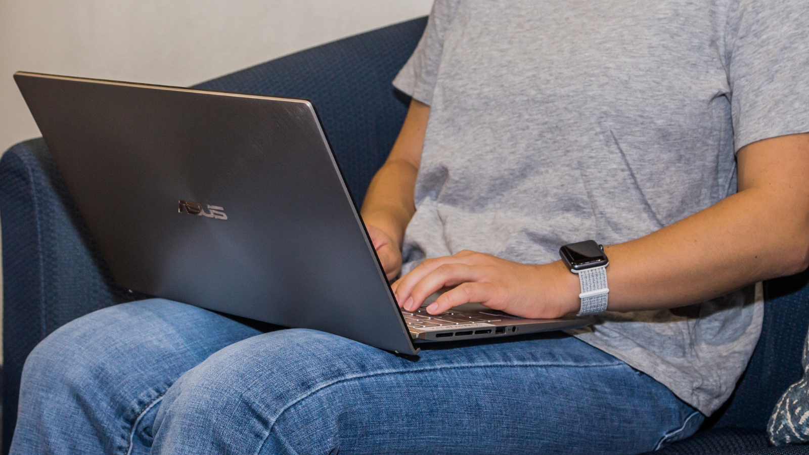 A person is typing on an ASUS laptop while sitting on a couch, wearing a gray t-shirt and a smartwatch.