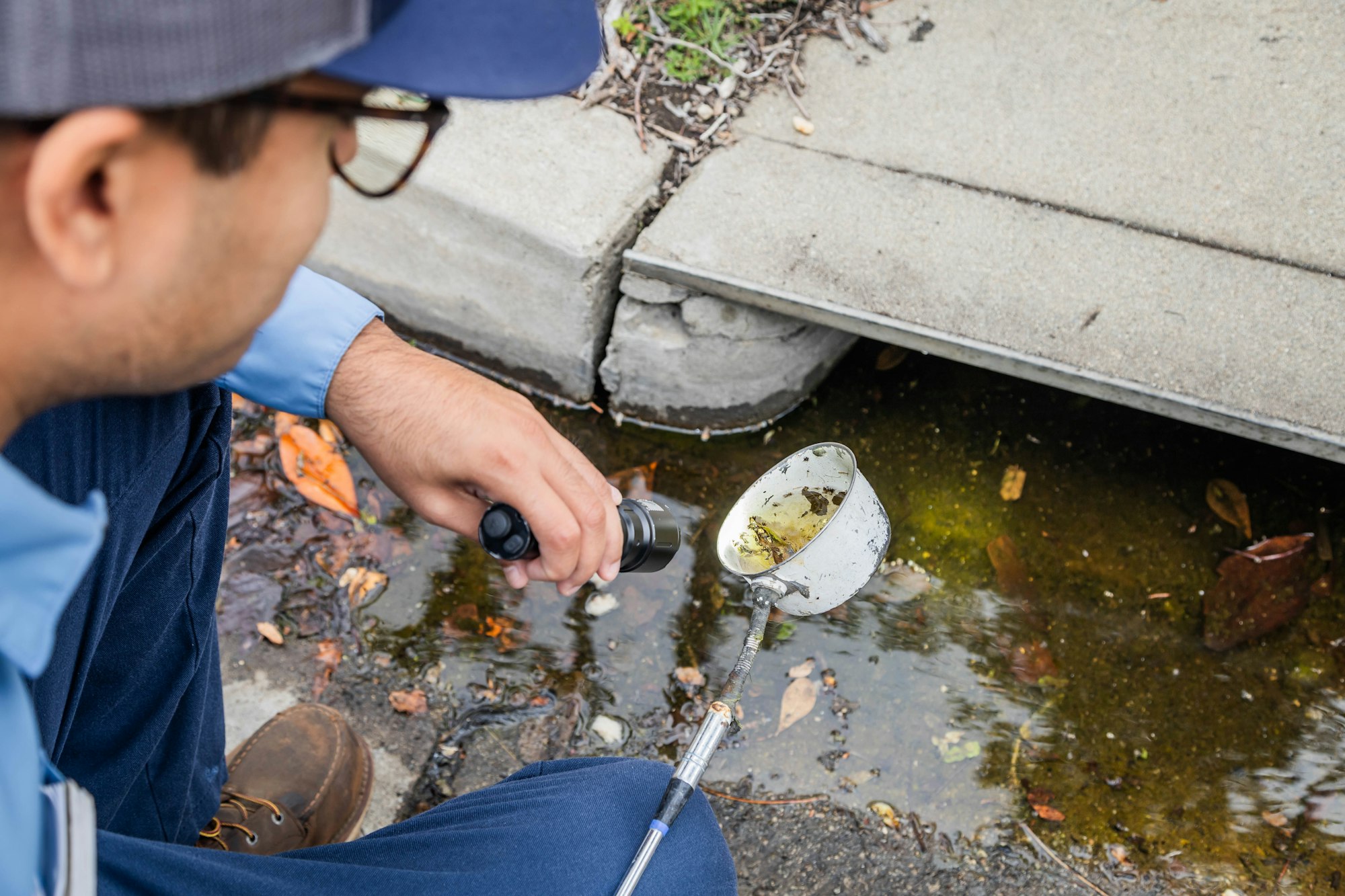 A person examines water in a drainage area using a handheld flashlight and a small cup. They appear to be conducting an inspection.