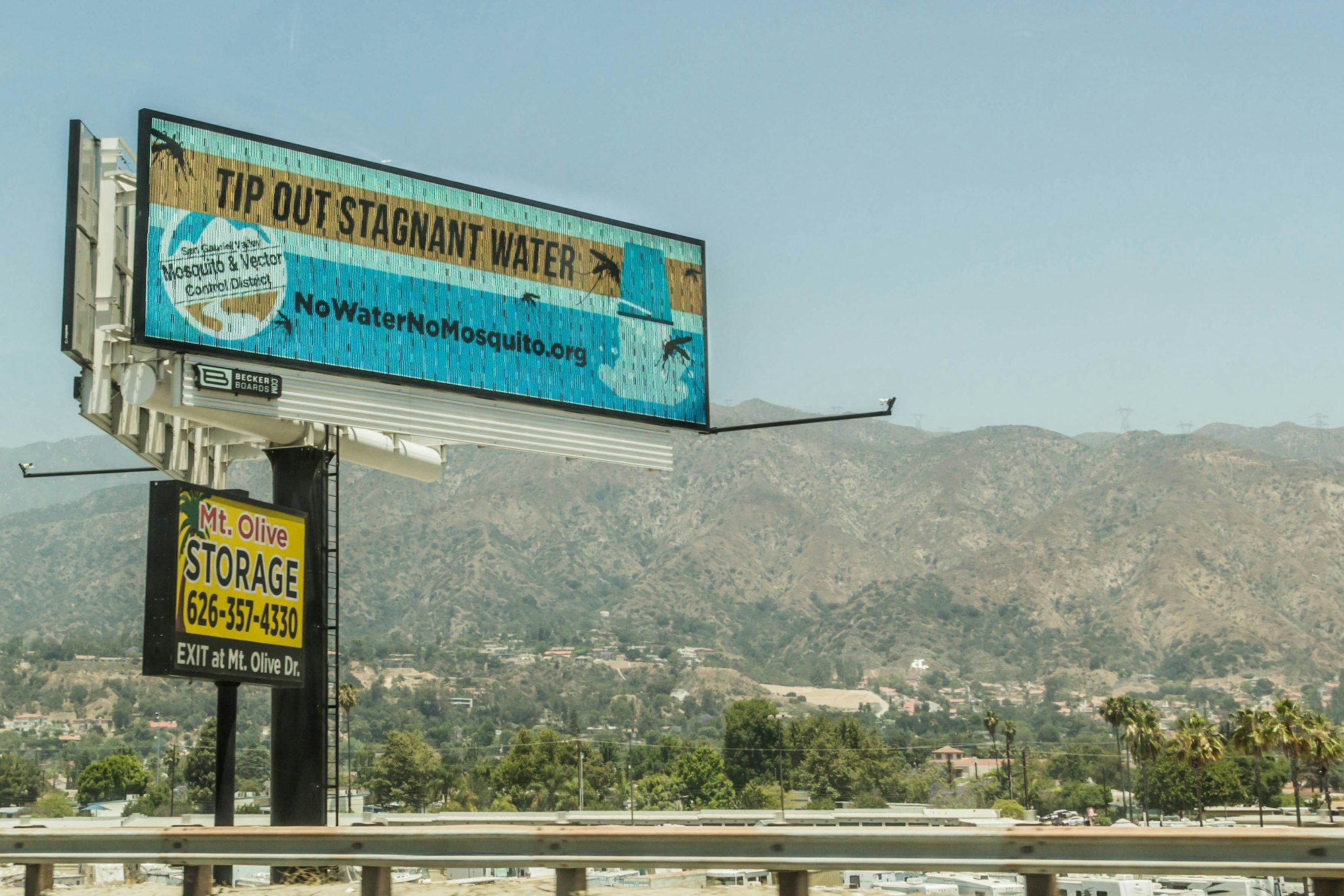 Billboard about preventing mosquito breeding, storage sign below, mountains in background, bright sunny day.