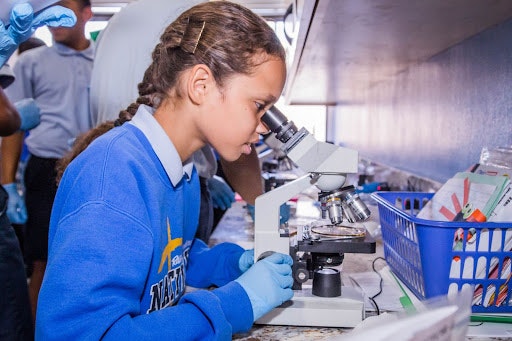 A girl in a blue sweater peers into a microscope while others observe, engaging in a scientific activity.