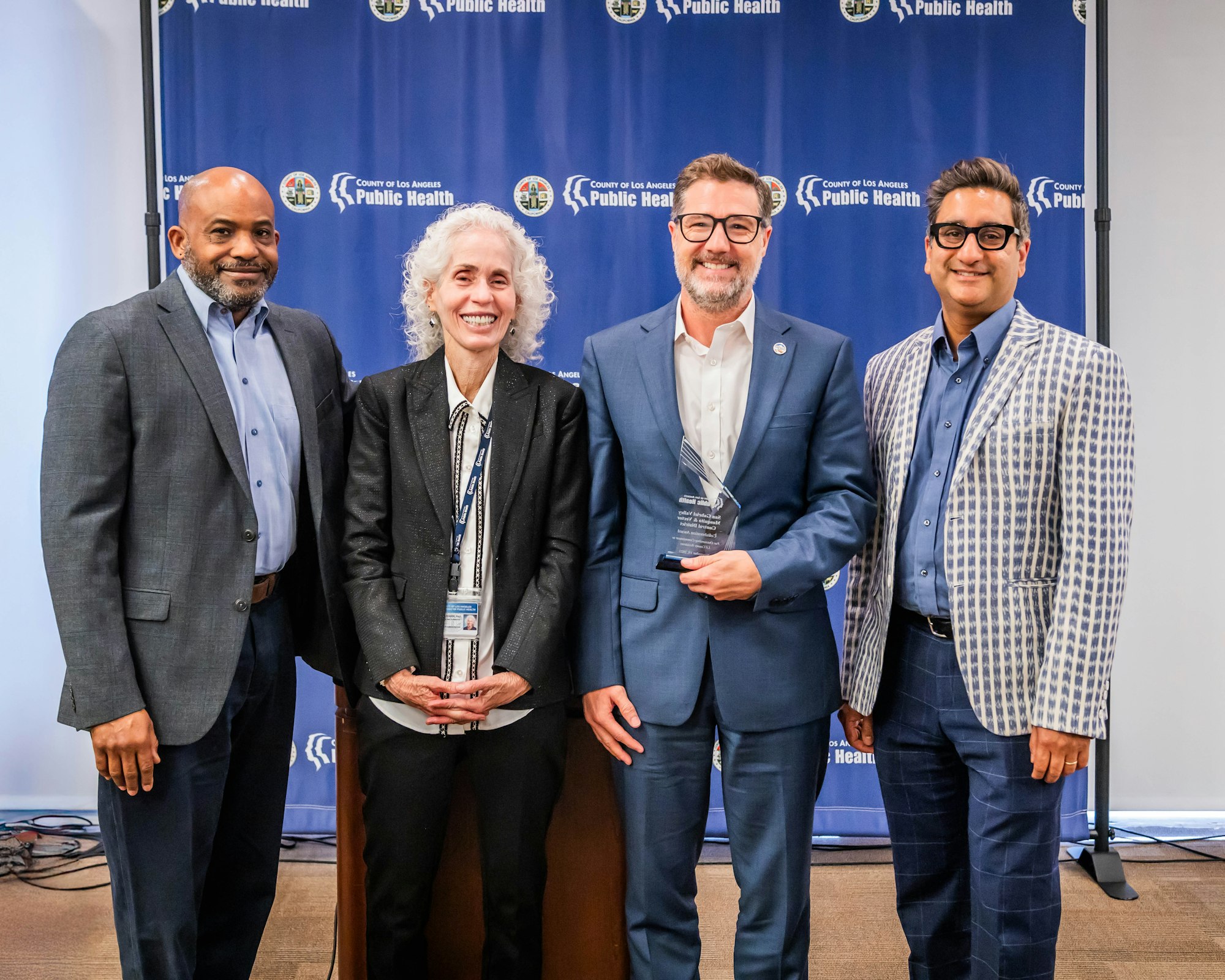A group of four individuals poses at a public health event, with awards and a backdrop displaying "County of Los Angeles Public Health."