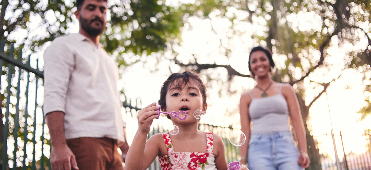 A child blows bubbles in a park, with two adults in the background, and trees around them.