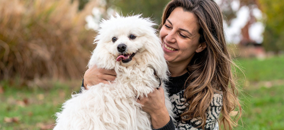 A woman happily embraces a fluffy white dog in a green outdoor setting, radiating joy and affection.