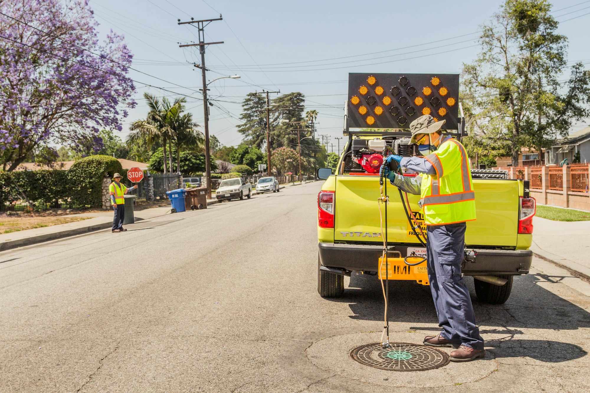 Road maintenance crew with equipment and a truck, one person holding a stop sign, in a sunny residential area.