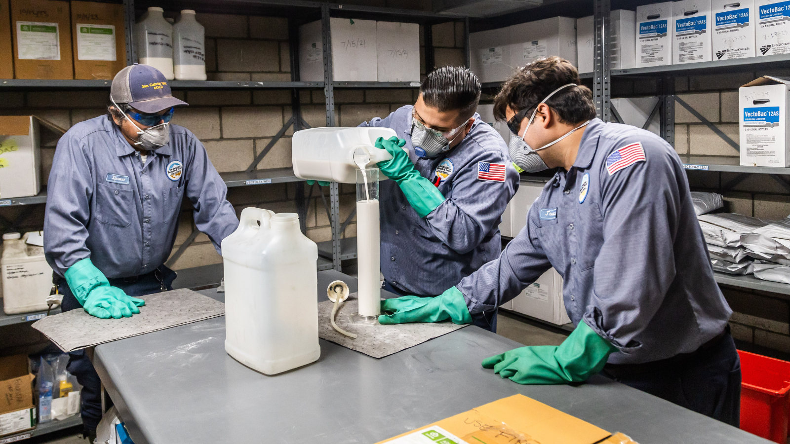 Three workers in protective gear are mixing chemicals in a laboratory setting, focused on their task.
