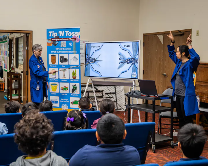 Two presenters discuss mosquito types and prevention strategies, engaging an audience of children during a workshop.
