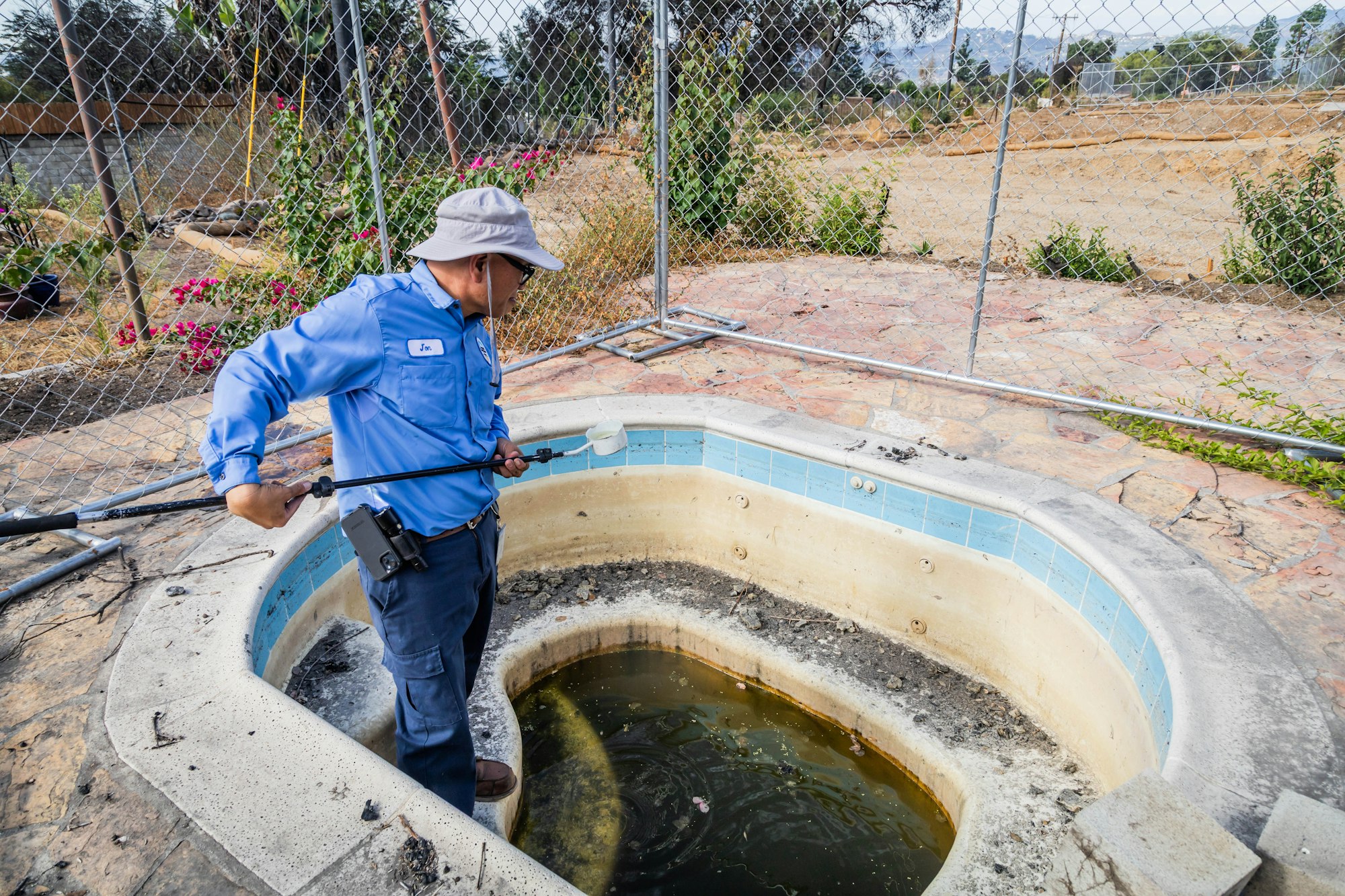 A person in a blue shirt inspects a dirty hot tub with a stick, surrounded by a fenced area and overgrown plants.