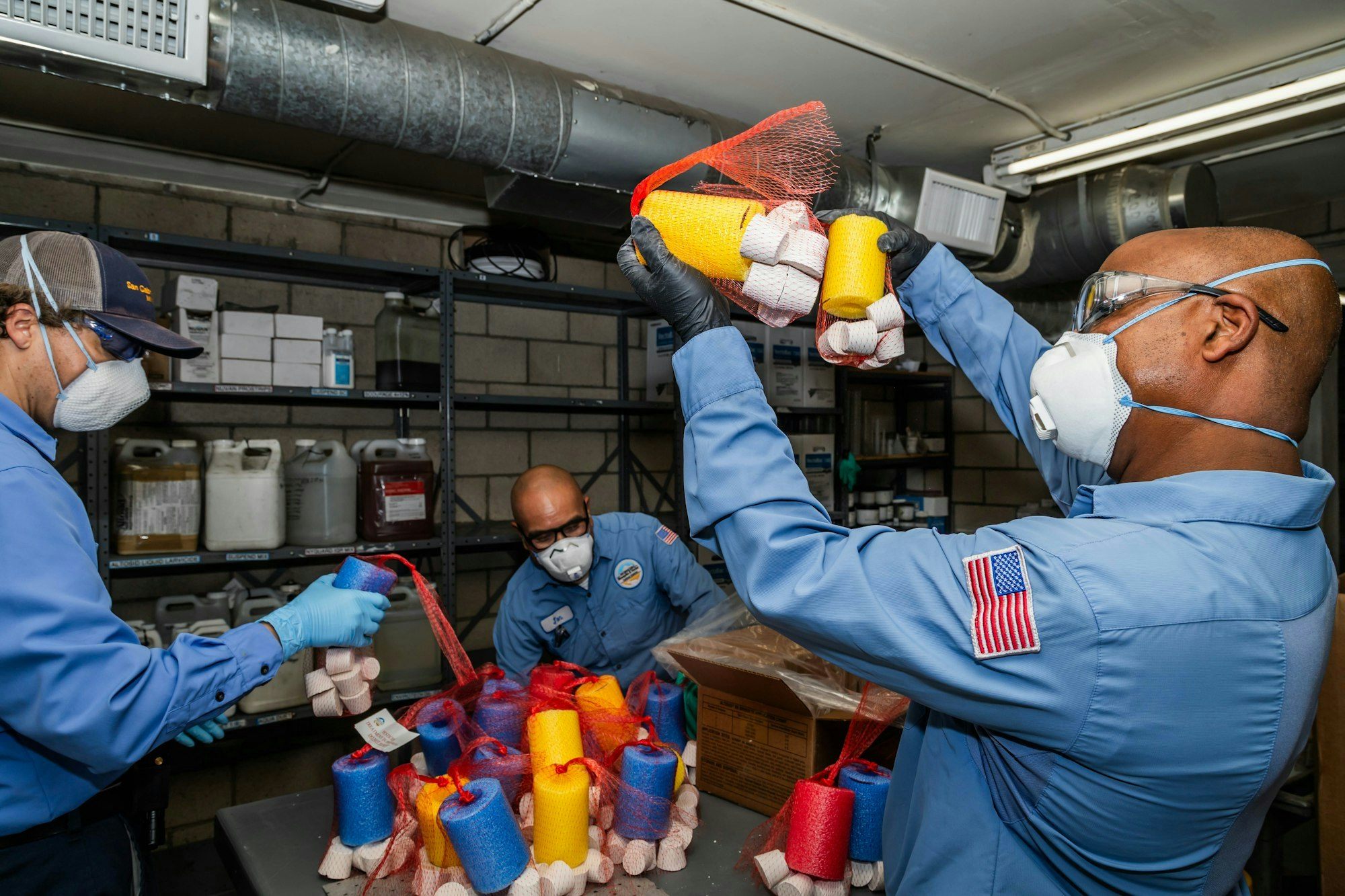 Workers in protective gear handle colorful items, possibly cleaning supplies, in a storage area with shelves and chemicals.